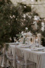 Elegant outdoor destination wedding table setup with white linens, floral centerpieces, transparent chairs, and string lights, creating a romantic and intimate atmosphere for a special celebration