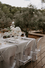 Elegant outdoor destination wedding table setup with white linens, floral centerpieces, transparent chairs, and string lights, creating a romantic and intimate atmosphere for a special celebration