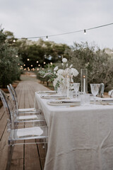 Elegant outdoor destination wedding table setup with white linens, floral centerpieces, transparent chairs, and string lights, creating a romantic and intimate atmosphere for a special celebration