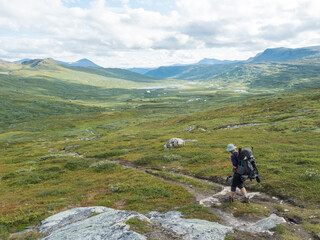 Fototapeta na wymiar Lonely man hiker figure with backpack and fishing rod at Lapland landscape with green valley, mountains and winding river stream. Wild nature at Padjelantaleden hiking trail. Summer day