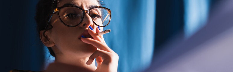 stylish woman in vintage eyeglasses smoking on blue background, banner