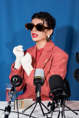 vintage style woman in sunglasses and white gloves talking during interview on blue background