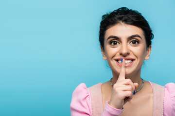 excited woman showing hush gesture while looking at camera isolated on blue