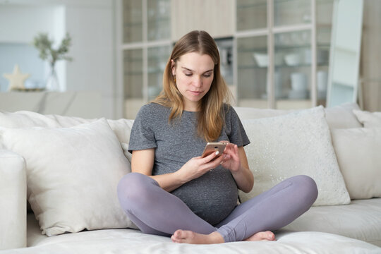 A pregnant woman is sitting on a sofa in the living room with a mobile phone in her hands.