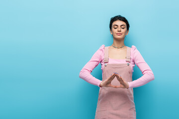 positive woman in pastel clothes relaxing in meditation pose isolated on blue