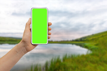 woman holding mobile phone with green display in hand against natural lake landscape