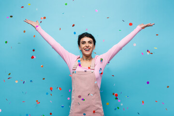 Obraz na płótnie Canvas excited woman in pastel clothes smiling under falling confetti on blue background