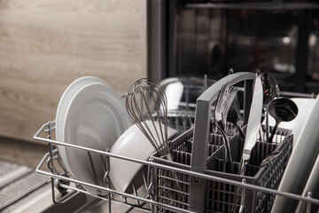 Front view of open automatic stainless steel built-in fully integrated top control dishwasher range machine with clean utensils, cutlery, glasses, dishes, plates inside in modern home kitchen