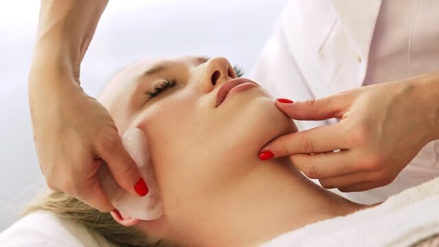 Beautician cosmetologist makes lifting modeling face massage with Gua Sha quartz scraper, professional cosmetic procedure in a beauty clinic salon for client. Skin care and cosmetology spa concept.
