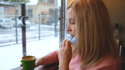 a blonde girl sits in a cafe opposite the window takes off his mask, takes a sip of coffee and puts...