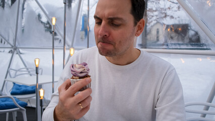 a man in a white sweater sits in the cafe tent sphere and eats a blueberry cupcake