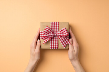 First person top view photo of valentine's day decorations woman's hands holding kraft paper giftbox with checkered ribbon bow on isolated beige background