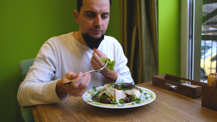 a man sits at a table in a restaurant wearing a mask, lowers the mask puts food in his mouth and puts the mask back