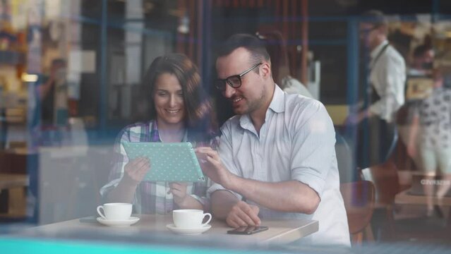 View though window of young couple use tablet sitting in coffee shop. Realtime