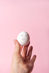Fototapeta na wymiar Easter holiday concept with cute handmade egg. Cute face egg in a female hand on a pink background