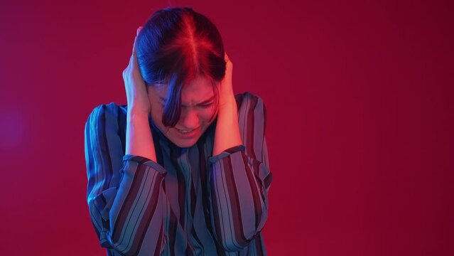 Loud noise. Annoying sound. Migraine problem. Irritated displeased woman plugging ears with fingers in neon light on dark red background.