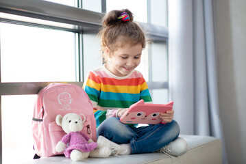 Positive little girl, holding tablet computer in her hands. Advertising new gadget, educational app. The girl sits near the window. She is wearing a colored T-shirt. Games.Children use technology.