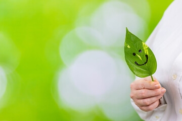 A woman holds a green leaf with a painted smile in her hands, a natural background with blurred greenery in the park, a creative idea, development in harmony with nature, work-life balance, copy space