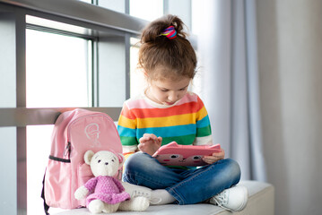 Positive little girl, holding tablet computer in her hands. Advertising new gadget, educational app. The girl sits near the window. She is wearing a colored T-shirt. Games.Children use technology.
