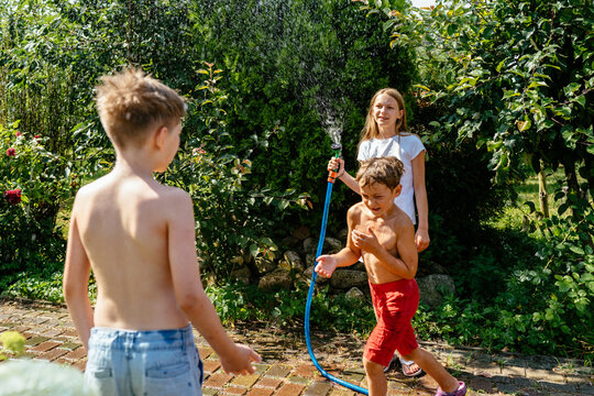 Cute blond girl is pouring a water from a hose at her brothers at backyard cozy garden at country house at summer day.