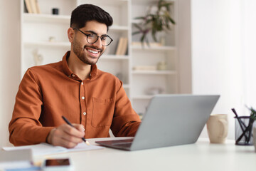 Smiling Arab man in glasses using laptop and writing