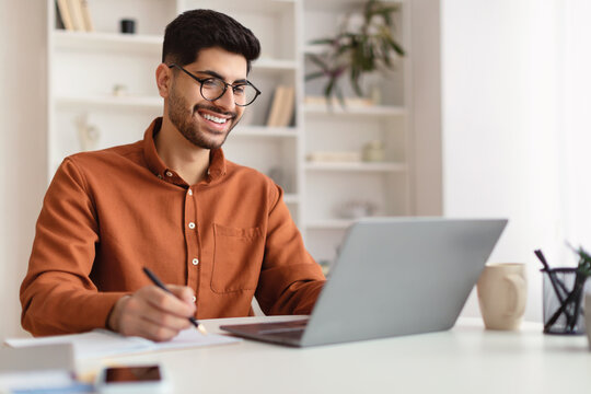 Smiling Arab man in glasses using laptop and writing