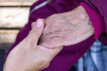 A mature woman holds her elderly mother's hand as they sit on a bench