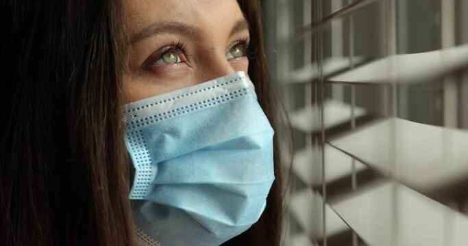 Woman wearing face mask looking out of window. Young ill woman looking sad with protective mask at home behind window in quarantine and lockdown during Covid