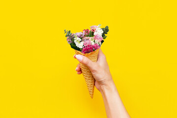 Ice cream waffle cone with flowers inside in a female hand isolated on a color yellow background....