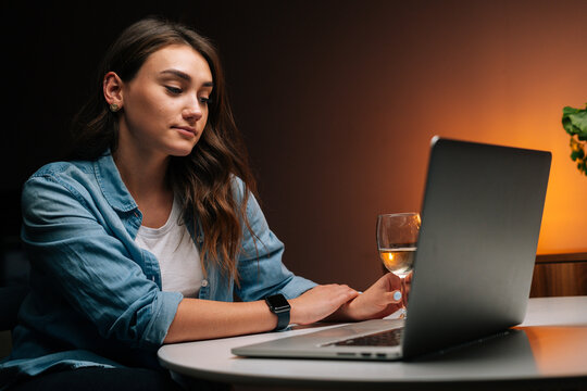 Close-up of pretty young woman using laptop sitting at table holding in hand glass with wine in cozy dark room. Tired female drinking alcohol during working on laptop at nighttime, looking on screen. - Powered by Adobe