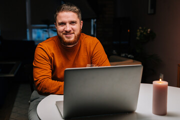 Smiling young man enjoying distant dating using laptop computer, holding glass with wine , talking celebration toasting via video call, sitting at table with candle in dark room, looking at camera.