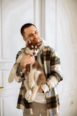 a young Caucasian man with dark hair in a plaid shirt plays with siberian husky dogs. The guy owner holding a husky puppy in a white room