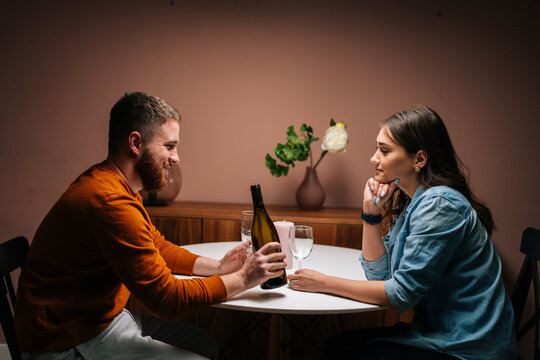 Side view of cheerful young man pouring champagne into glass during talking with wife sitting together at festive table. Love couple celebrating anniversary or Valentine day having romantic dinner