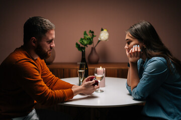 Side view of handsome loving young man making proposal for marriage to charming amazed woman at table with candles, marry me. Beautiful romantic couple having dinner celebrating Valentines day.