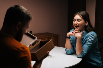 Happy young man opening box with received gift from cheerful wife enjoying romantic dinner date on birthday or Valentines Day. Pretty young woman giving wrapped box with gift to husband at home.