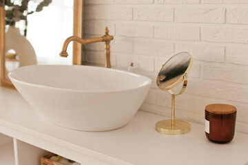 Bathroom decor with a mirror and candle and sink, beautiful corner for daily beauty routine. Modern bathroom interior design with gold elements and details and predominantly in white color.