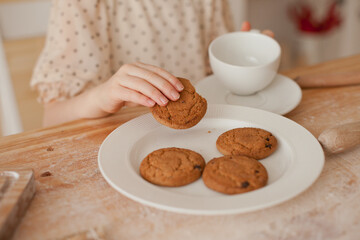 Little girl eating freshly baked cookies in the kitchen table covered with flour after baking. Kid eating tasty cookies from white plate.