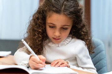 Serious little multiracial curly girl having distant education while writing something
