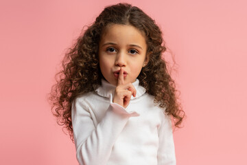Portrait of adorable little girl making silence gesture with finger on lips, looking at the camera