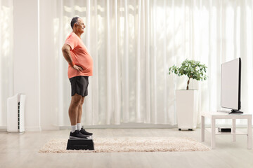 Fototapeta na wymiar Full length profile shot of a mature man in sportswear standing on a stepper in front of tv