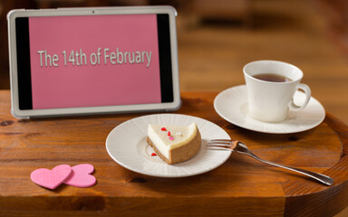A Valentine's Day card, a piece of cheesecake with a mug of tea and two pink hearts on a wooden table and a tablet with the inscription 