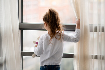 Little girl stands near a window with teddy bear in her hands. She is dressed in a white jumper. Childhood. Lifestile.