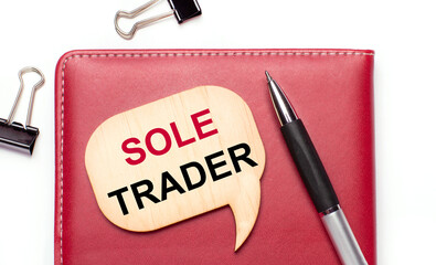 On a light background there are black paper clips, a pen, a burgundy notepad a wooden board with the text SOLE TRADER