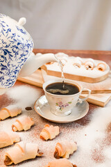 The teapot pours boiling water into a cup of coffee against the background of croissants with apple jam sprinkled with powdered sugar.