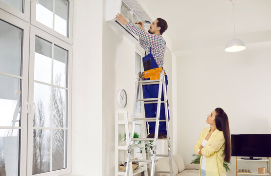 Technician Repairing AC At Work. Repair Service Guy Standing On Ladder In Young Lady's Home And Opening Up Modern White Wall Mounted Air Conditioner In Order To Check, Do Disinfection Or Fix Troubles