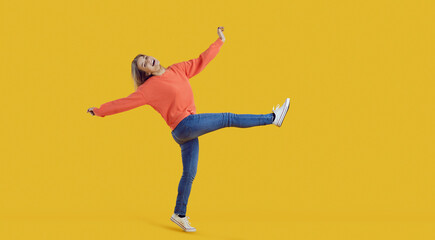 Fototapeta na wymiar Funny carefree young woman in comfortable casual wear having fun and fooling around. Happy pretty teenage girl in orange sweatshirt and blue jeans dancing in studio with vibrant yellow background