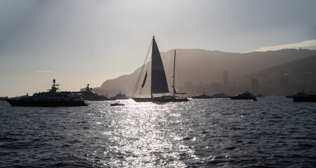 A lot of huge sail yachts and motor boats in port of Monaco at sunset, mountains are on background, megayachts are moored in sea, sun reflection on water, backlight