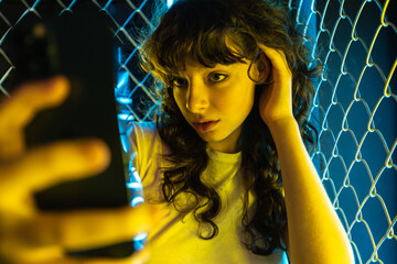 A young woman with curly hair takes a selfie near a yellow metal grid and in neon lighting.