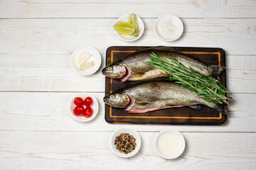 Raw fish, trout with on cutting board with ingredients for cooking. Fish, pepper, garlics, tomatoes, lemon and rosemary on white wooden table. Healthy food and dieting concept flat lay, copy space.