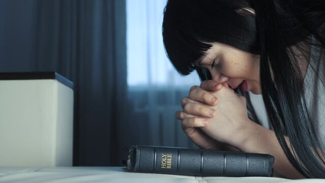 girl praying indoors at bedtime on bible. lifestyle religion concept evening prayer believer woman brunette hands on bible praying by bed